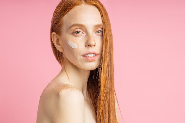 Beautiful young ginger woman with clean perfect skin. Portrait of happy beauty model with bare shoulders, no makeup, applying face cream, smiling against pink wall. Spa, skin care and wellness.