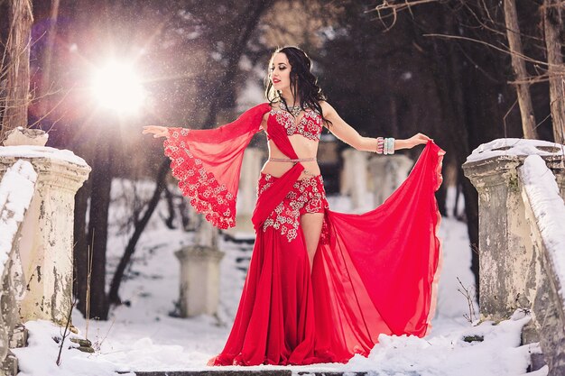 Beautiful young girl dancing belly dance in red dress in winter in a park on the snow.