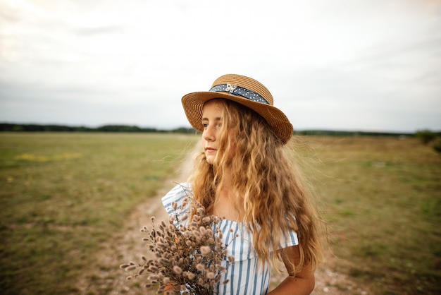 Beautiful young girl in a dress with blonde hair and a hat walks across the meadow