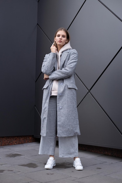 Beautiful young girl in fashionable biege hoodie elegant gray coat and pants on dark silver background Walking on the street