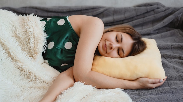 Beautiful young girl in a green nightie sleeps on a yellow pillow on a sofa under a white blanket