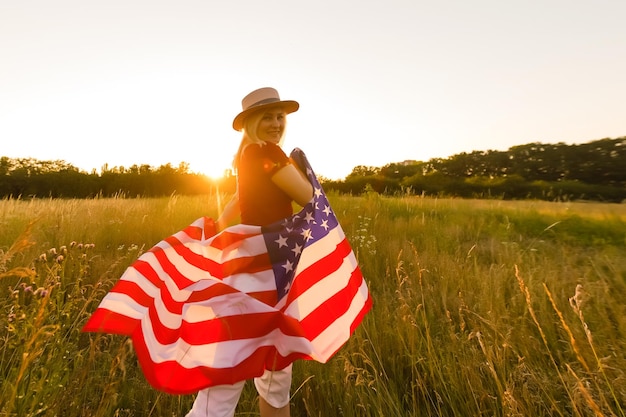 Beautiful young girl holding an American flag in the wind in a field of rye. Summer landscape against the blue sky. Horizontal orientation.