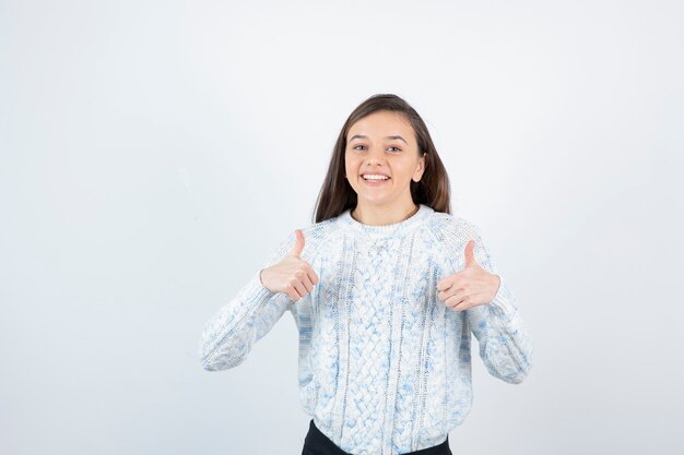 Beautiful young girl in knitted sweater standing and giving thumbs up.