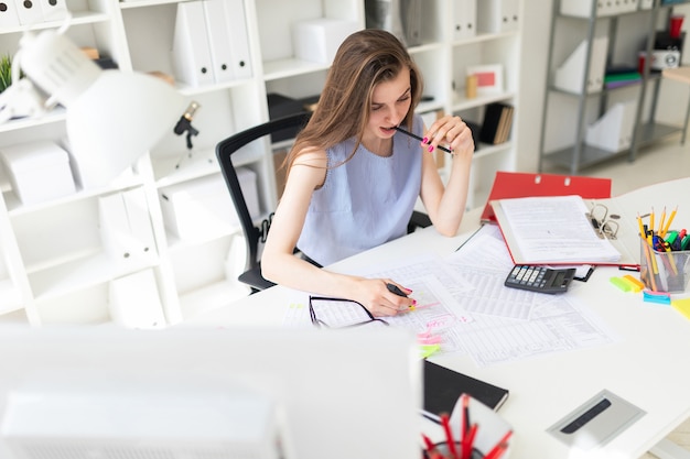 Beautiful young girl in the office sits at a table, is holding a yellow marker in her hand and gnawing a pencil.