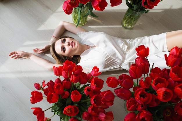 A beautiful young girl in a peignoir is lying on the floor between large bouquets of tulips March 8 concept