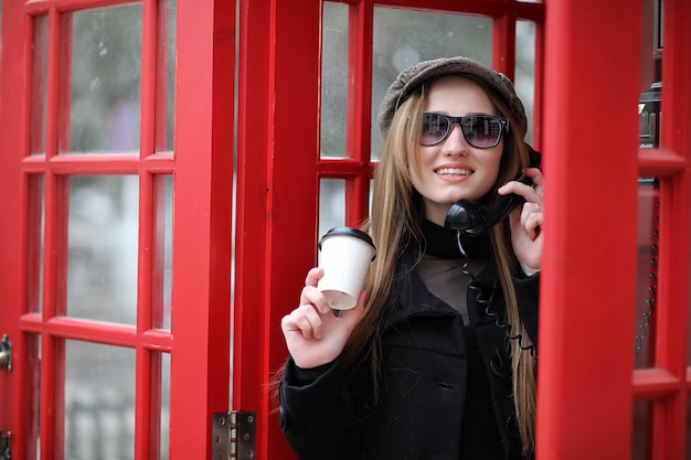 Beautiful young girl in a phone booth. The girl is talking on the phone from the payphone. English telephone booth in the street and a woman talking on the phone.