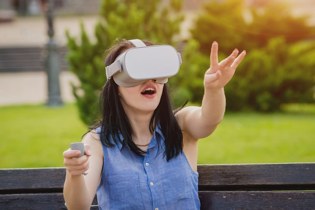 A beautiful young girl plays a game wearing virtual reality glasses on the street.