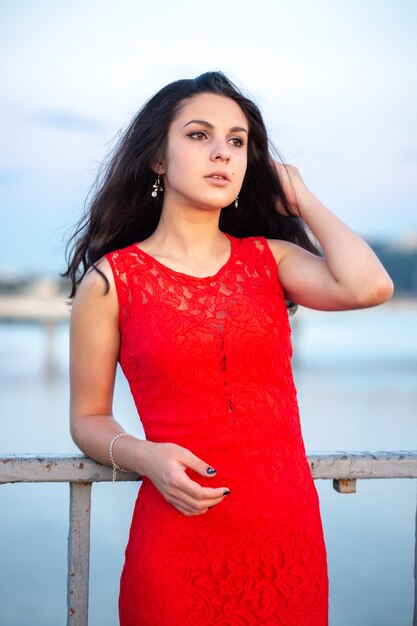 Beautiful young girl in a red dress posing on a bridge near an old fence. In the background there is a river, bridges and fragments of city buildings in the defocus.