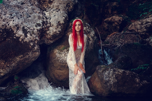 Beautiful young girl resting in water. Young woman in the white dress is sitting on the stone in the middle of a creek.