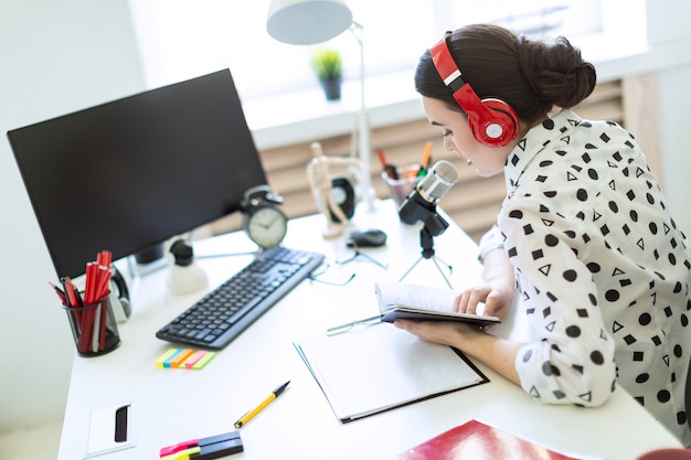 Beautiful young girl sits in headphones and with a notepad at the table in the office and speaks into the microphone.