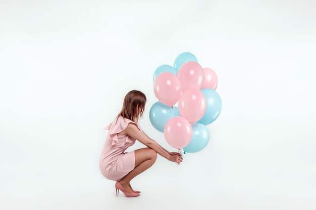 A beautiful young girl sits with blue and pink balloons on a white wall.