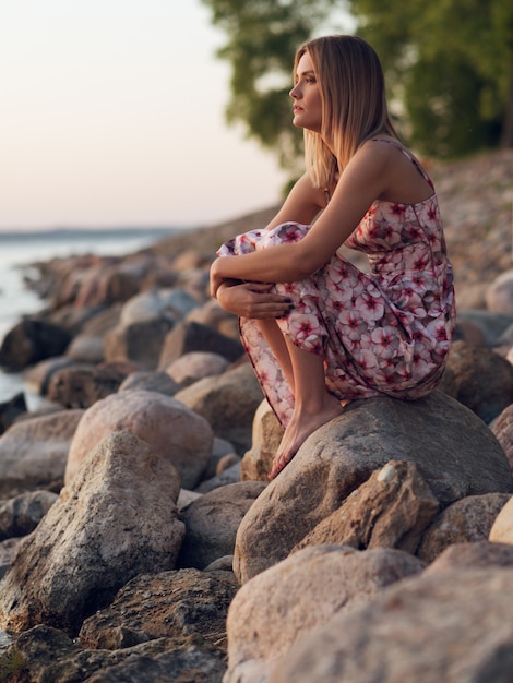 Beautiful young girl sitting on the rocky shore in a dress and looking at the sunset