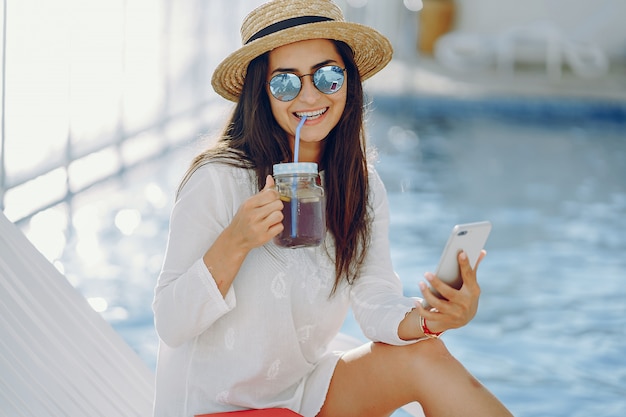 A beautiful young girl sitting on a summer terrace by the pool with phone and cocktail