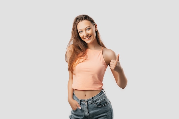 Beautiful young girl smiling and shows thumbs up gesture hand on a white isolated background. Positive woman points to an idea, a place for advertising