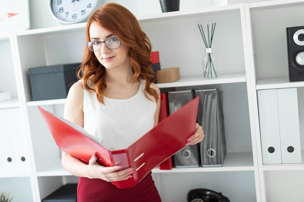 A beautiful young girl stands near a stack in the office and holds a folder with documents in her hands.