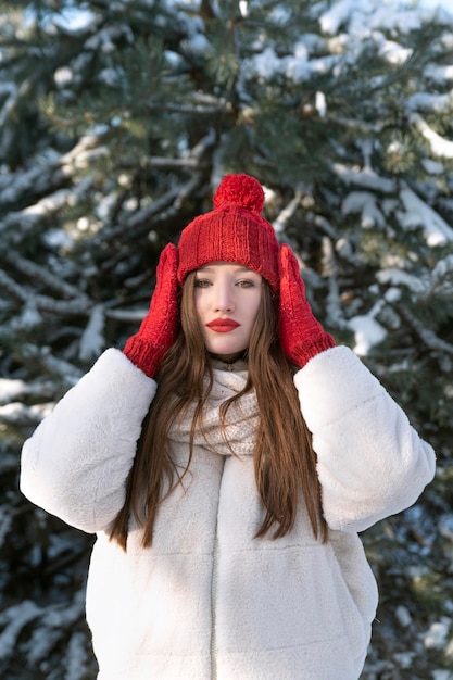 Beautiful young girl straightens hat in winter park against snowy pine trees background Vertical frameSunny winter day