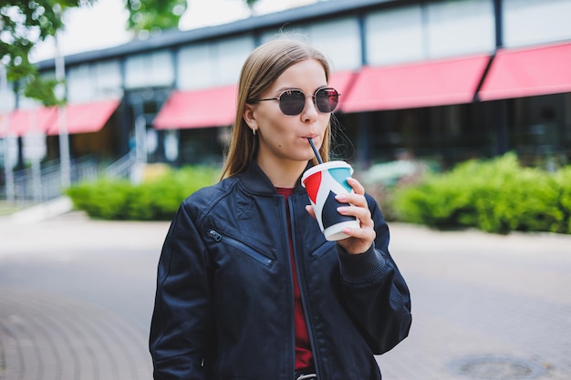 Beautiful and young girl in sunglasses writes a message on a cell phone and drinking coffee outdoors