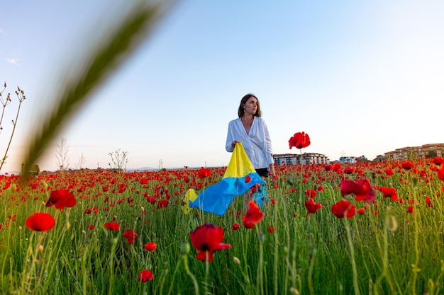 A beautiful young girl in a white shirt and shorts poses in a poppy field with the flag of Ukraine