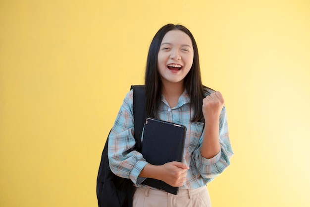 Beautiful young girl with backpack and hold laptop on yellow background.