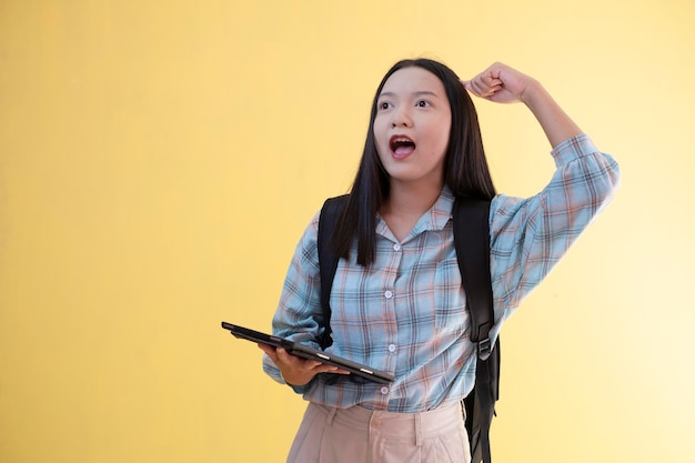 Beautiful young girl with backpack and hold laptop on yellow background.