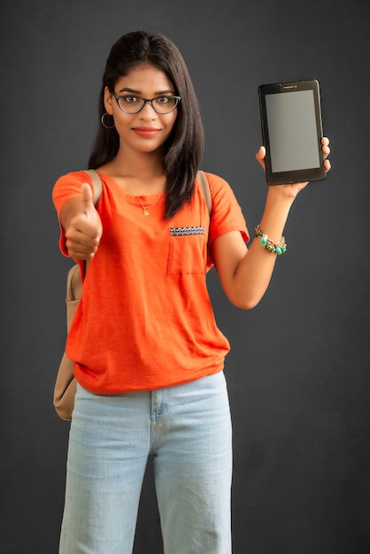 A beautiful young girl with a backpack shows a blank screen of a smartphone or mobile or tablet phone posing on a grey background