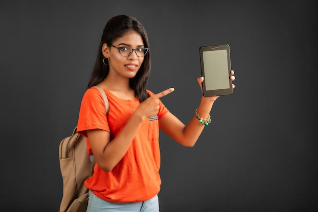 A beautiful young girl with a backpack shows a blank screen of a smartphone or mobile or tablet phone posing on a grey background