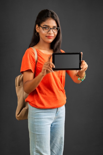 A beautiful young girl with a backpack shows a blank screen of a smartphone or mobile or tablet phone posing on a grey background