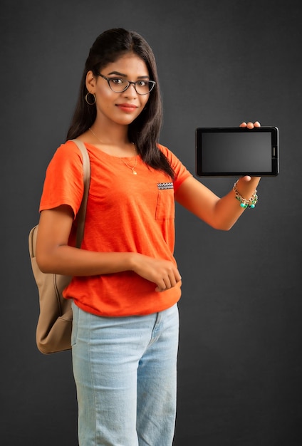 A beautiful young girl with a backpack shows a blank screen of a smartphone or mobile or tablet phone posing on a grey background