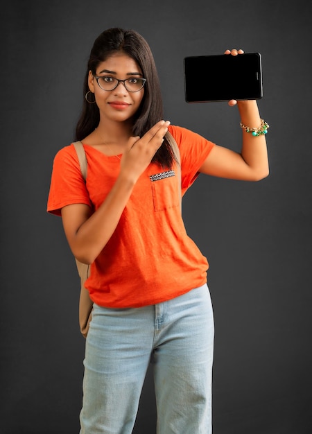 A beautiful young girl with a backpack shows a blank screen of a smartphone or mobile or tablet phone posing on a grey background