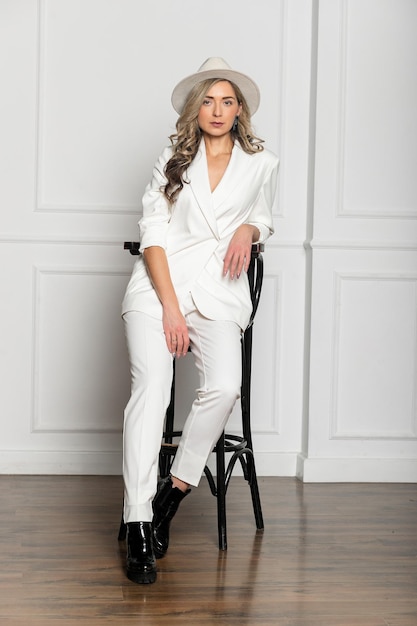 Beautiful young girl with a beautiful figure in a white trouser suit and a widebrimmed white hat posing against a white background in the studio sitting on a chair