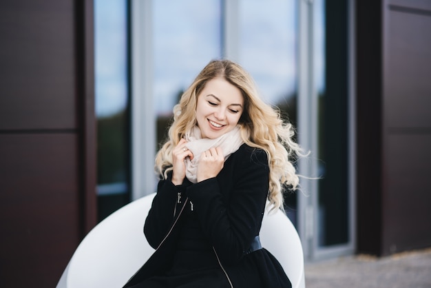 Beautiful young girl with blond wavy hair in a black coat sits on a modern building