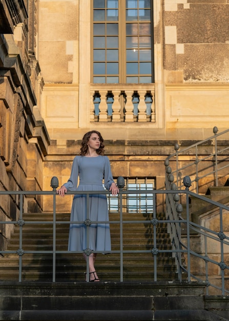 A beautiful young girl with dark hair in a gray dress stands on the carved stairs of the palace Grace and elegancesetting sun