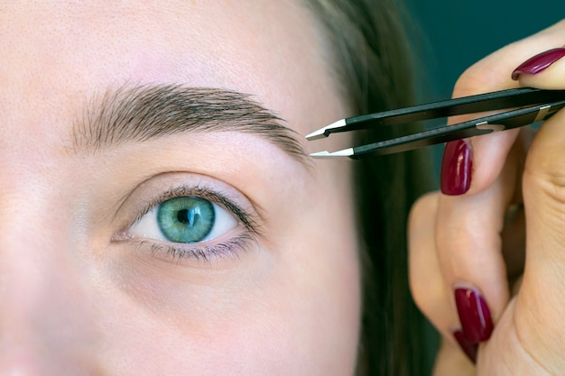 Beautiful young girl with eyelashes tweezing her eyebrows in a beauty salon. Woman doing eyebrow permanent makeup correction . Microblading brow.