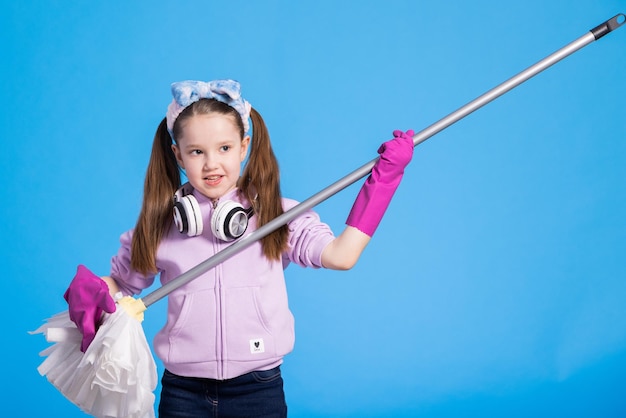 Beautiful young girl with headphones and mop sings on blue background pretending to play guitar