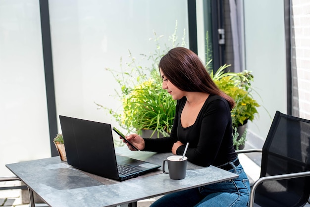 A beautiful young girl with a laptop sits at a table and holds a mobile phone in her hands