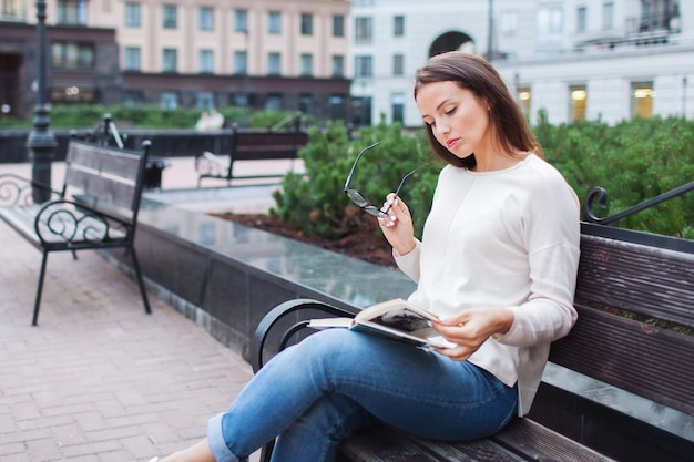 A beautiful young girl with long brown hair sitting on a bench with a book