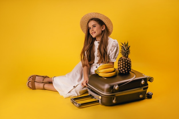 A beautiful young girl with long hair has planned a trip and sits near a suitcase with fruits