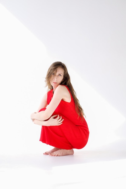 Beautiful and young girl with long hair in a red dress sits on the floor of a cyclorama and poses