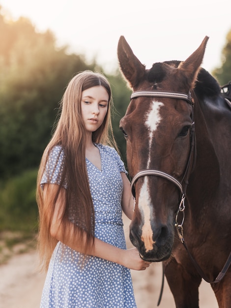 A beautiful young girl with long hair stands next to a horse in nature in the summer