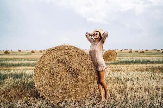 Beautiful young girl with long hair in sunnglasses and straw hat posing on a wheat field near hay bales. Happy brunette in summer dress