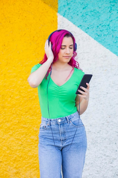 Beautiful young girl with pink hair listening to music on headphones street style colorful wall ba