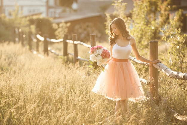 Beautiful young girl with pions on the nature otdoors in the sunshine. Pretty woman outside with flowers in the pink skirt