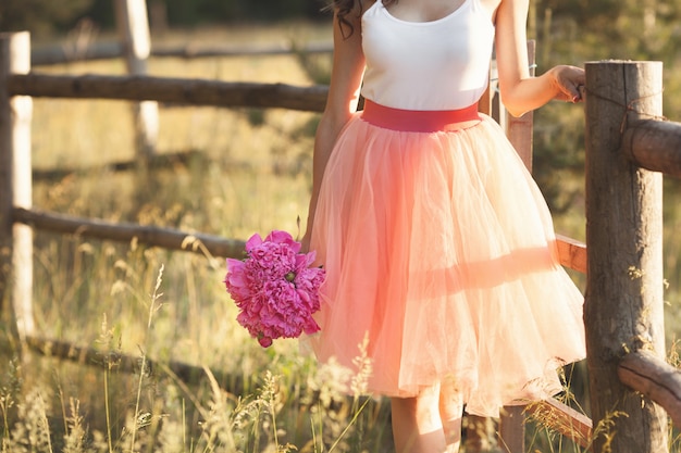 Beautiful young girl with pions on the nature outdoors in the sunshine. Pretty woman outside with flowers in the pink skirt