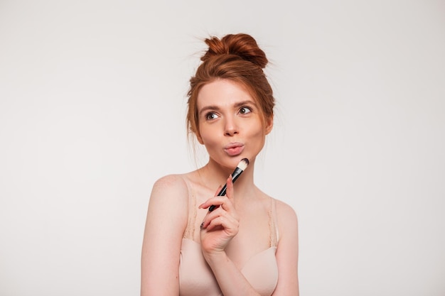 Beautiful young happy funny woman model with natural red hair holding a makeup brush and posing on a white background in the studio Beauty skin care and makeup concept