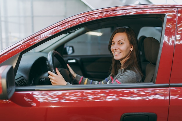 A beautiful young happy smiling European brown-haired woman with healthy clean skin dressed in a striped t-shirt sits in her red car with black interior. Traveling and driving concept.