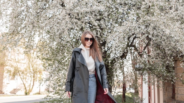 Beautiful young happy smiling fashion model woman with cool sunglasses in a fashionable leather long coat with stylish hoodies and vintage jeans walks in the city near a flowering tree