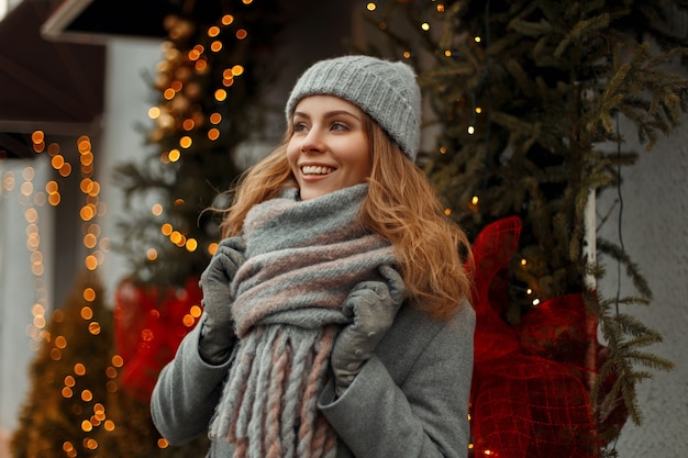 Beautiful young happy woman with a magic smile in fashion knitwear clothes in a knitted gray hat and coat with a stylish scarf on the street near the lights and Christmas garlands