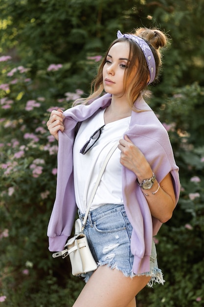 Beautiful young hipster girl with bandana in trendy casual clothes: purple sweatshirt, ripped denim shorts and white t-shirt with handbag and women's accessories outdoors