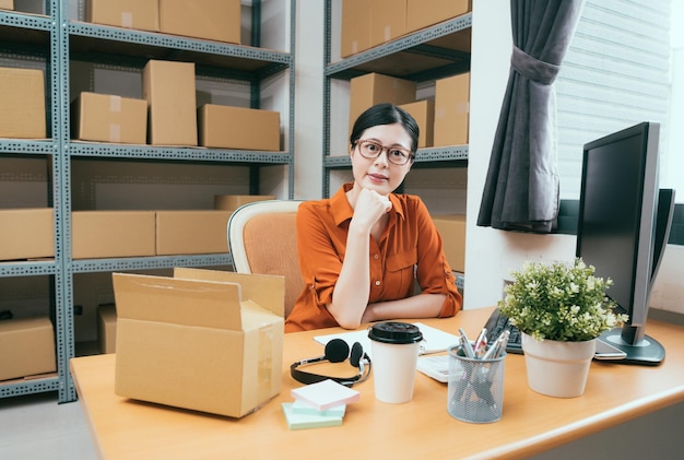 beautiful young lady as online shopping owner sitting on working desk doing order data and face to camera at stock warehouse.