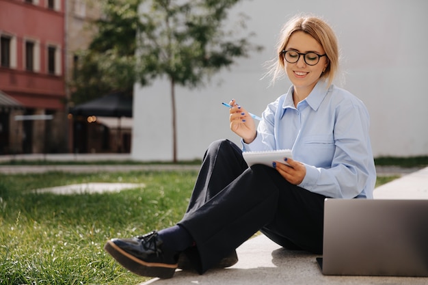 Beautiful young lady in eyeglasses and casual clothing studying on laptop at urban space. Self education and knowledge. Student spending free time outdoors.